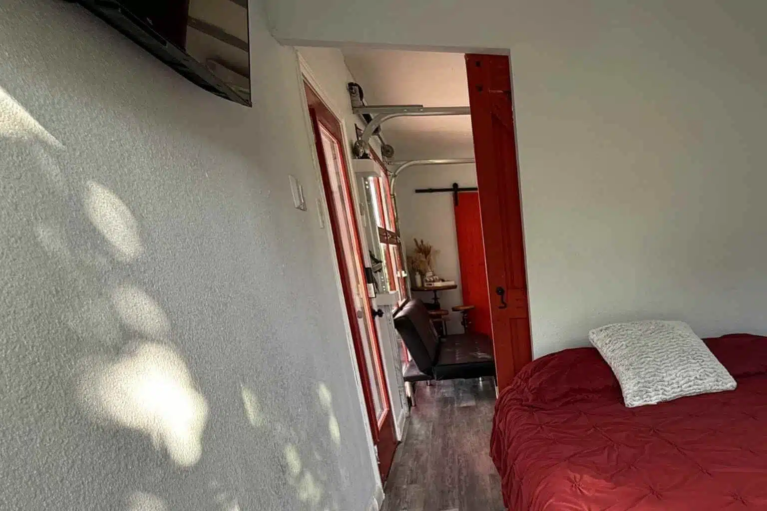 A room with a red bed, white pillow, and open doorway showing a chair and another room.