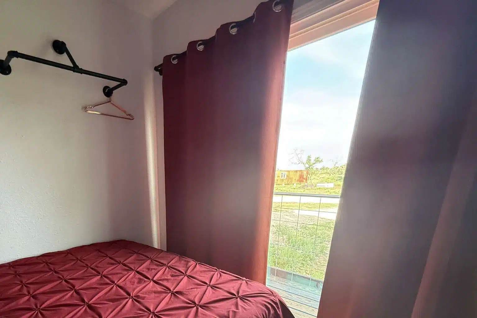 A room with a red bedspread, a window with curtains, and a view of grass outside.